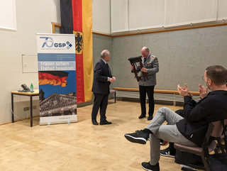 Ein Offizier lächelt, während er einen kleinen Klapphocker von einem Mann im Anzug erhält, mit einem GSP-Banner im Hintergrund.
