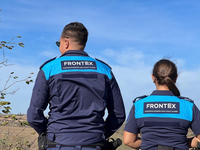 Zwei Frontex-Beamte der Europäischen Grenz- und Küstenwache in Uniform, von hinten gesehen, blicken unter blauem Himmel hinaus.