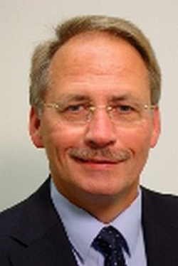 Professionelles Porträt eines Mannes mittleren Alters mit hellem Haar, Brille, Schnurrbart und dunklem Anzug.