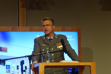 Ein Bundeswehr-Offizier, André Wüstner, spricht an einem Pult mit Mikrofon, im Hintergrund ein blauer Bildschirm mit Wappen.