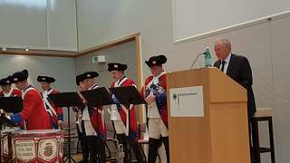 Eine historische Bürgergarde-Kapelle in roten Uniformen spielt Instrumente, während ein Mann am Bundessprachenamt-Rednerpult spricht.