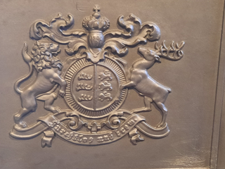 Geprägtes königliches Metallwappen: gekrönter Schild, gestützt von Löwe und Hirsch, mit Spruch 'Gott mit uns'.