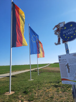 Deutsche und EU-Flaggen wehen neben einem 'DEUTSCH-LAND' Schild auf einem grünen Feld mit Weg unter blauem Himmel.