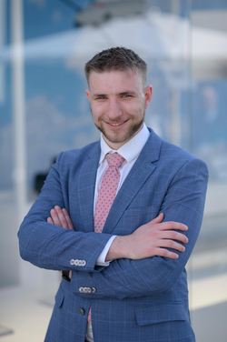 Junger Geschäftsmann in einem blauen Anzug und rosa Krawatte, lächelnd mit verschränkten Armen, verschwommener blauer Hintergrund.