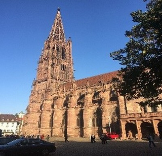 Freiburger Münster, eine gotische Kathedrale, mit ihrem aufwendigen Turm vor blauem Himmel. Menschen und Autos auf dem Platz.