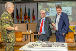 Drei Männer, darunter ein Soldat in Uniform, diskutieren um ein Architekturmodell eines Gebäudekomplexes. Im Hintergrund sind Flaggen und eine Präsentation sichtbar.