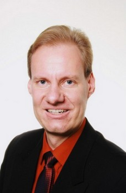 Kopfaufnahme eines lächelnden Mannes mit kurzen braunen Haaren, dunklem Anzug und rotem Hemd.