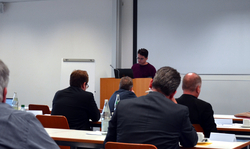 Ein Redner an einem Pult mit Laptop spricht ein Publikum an, das an Tischen in einem hellen Konferenzraum sitzt.