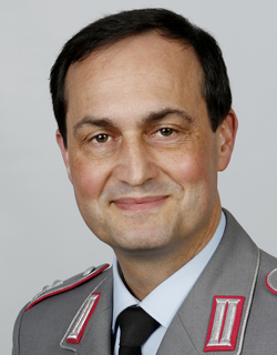 Porträt eines lächelnden Mannes in grauer Militäruniform mit roten und silbernen Abzeichen.