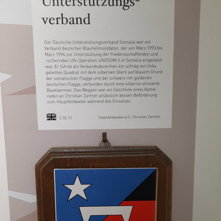 Holzplakette mit dem UNOSOM II Wappen, das den somalischen Stern auf Blau und die deutschen Flaggenfarben mit weißer Klammer zeigt.