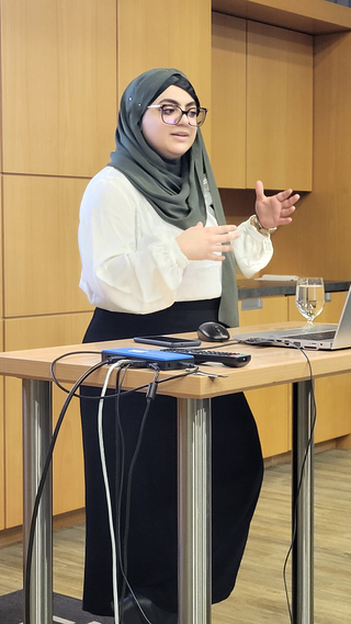 Eine Frau mit grünem Hijab und Brille steht hinter einem Tisch mit Laptop, Handy und Wasser und gestikuliert.