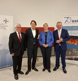 Vier Personen, darunter eine Frau, stehen lächelnd vor Bannern der Deutschen Atlantischen Gesellschaft und der GSP, mit lettischer Flagge.