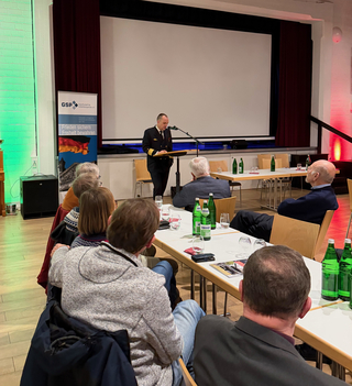 Ein Marineoffizier spricht vor einem Publikum mit einem GSP-Banner und großem Projektionsschirm im Hintergrund.