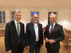 Drei lächelnde Männer, zwei in Anzügen und einer in Blazer und Jeans, stehen zusammen in einem Innenraum.