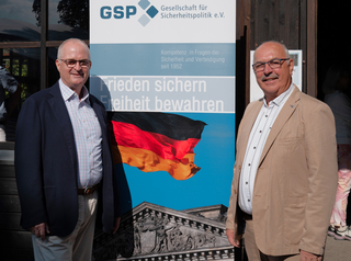 Zwei Männer lächeln vor einem GSP-Banner mit deutscher Flagge und Reichstagsgebäude.