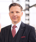 Porträt eines lächelnden Mannes mittleren Alters im dunkelgrauen Anzug mit roter Krawatte und blauen Augen.