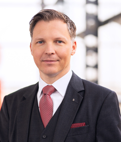 Porträt eines lächelnden Mannes mittleren Alters im dunkelgrauen Anzug mit roter Krawatte und blauen Augen.