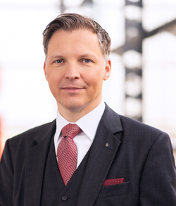 Porträt eines lächelnden Mannes mittleren Alters im dunkelgrauen Anzug mit roter Krawatte und blauen Augen.