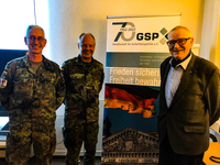 Drei lächelnde Männer, zwei in militärischen Tarnuniformen und einer in Zivilkleidung, vor einem GSP-Banner mit deutscher Flagge.