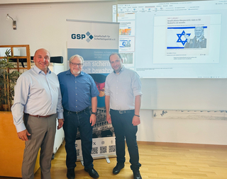 Drei lächelnde Männer stehen in einem Raum mit einem GSP-Banner und einem projizierten Nachrichtenartikel.