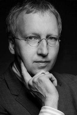 Schwarz-Weiß-Porträt eines Mannes mit runder Brille und Anzug, die Hand am Kinn.