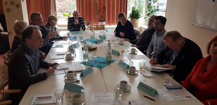 Eine Gruppe von Erwachsenen sitzt an einem Konferenztisch mit Getränken und Dokumenten, während eines Meetings.
