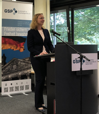 Lächelnde Frau mit Brille spricht an einem Rednerpult. Dahinter ein GSP-Banner mit Deutschlandfahne und Reichstagsgebäude.