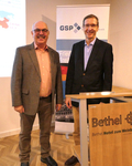 Zwei lächelnde Männer in Anzügen stehen vor einer Projektion mit einer Weltkarte und Bannern von GSP und Bethel.