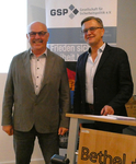 Zwei lächelnde Männer, einer im grauen, einer im dunklen Sakko, stehen vor einem GSP-Banner.