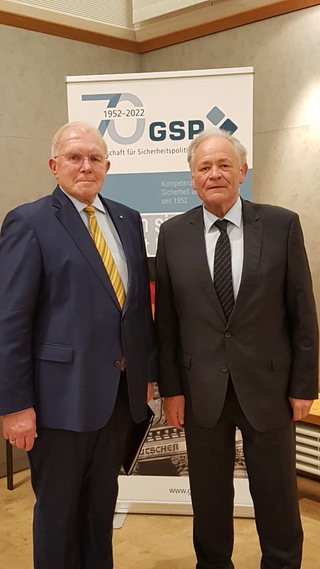 Porträt von zwei älteren Männern in Anzügen vor einem GSP-Banner zum 70-jährigen Jubiläum (1952-2022).