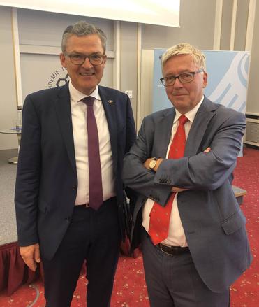 Zwei Männer in Anzügen und Brillen lächeln in die Kamera. Der linke trägt eine lila Krawatte, der rechte eine rote mit Muster.
