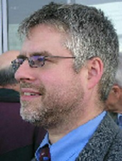 Porträt eines Mannes mittleren Alters mit Brille, graumeliertem Haar und Bart, im Profil nach links blickend.