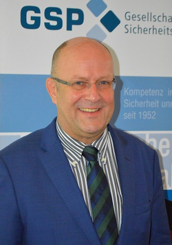 Porträt eines lächelnden, glatzköpfigen Mannes mit Brille, gestreiftem Hemd, Krawatte und blauem Anzug vor GSP-Hintergrund.
