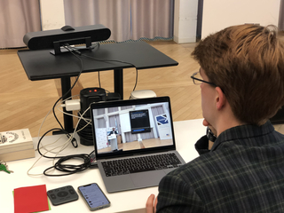 Person in kariertem Hemd mit Brille verfolgt virtuelle Konferenz auf Laptop, zeigt Redner und CASSIS-Logo.