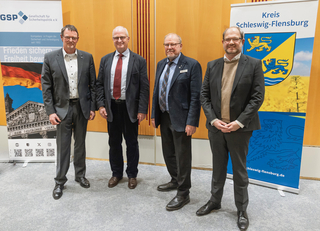 Vier lächelnde Männer in Anzügen vor Bannern der Gesellschaft für Sicherheitspolitik und Kreis Schleswig-Flensburg.