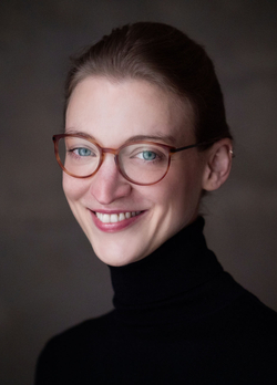 Porträt einer lächelnden Frau mit blauen Augen und brauner Brille in einem schwarzen Rollkragenpullover vor dunklem Hintergrund.