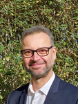 Ein lächelnder Mann mittleren Alters mit Brille und Bart im Anzug vor einer grünen Hecke.