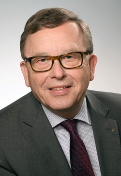 Kopfaufnahme eines lächelnden Mannes mit dunklem Anzug, hellem Hemd, roter Krawatte und brauner Brille.