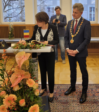 Eine Frau unterschreibt ein Dokument an einem blumengeschmückten Tisch, während ein Mann mit Amtskette zusieht.