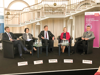 Podiumsdiskussion mit fünf Sprechern, darunter ein Militär, auf einer Bühne vor einer großen Treppe. Namen: Cremer, Shino, Wagner, Linntam, Bohnert.