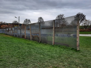Ein langer, verwitterter Zaun aus Betonpfosten und Wellblechpaneelen erstreckt sich über eine grüne Wiese unter grauem Himmel.