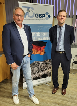 Zwei Männer lächeln vor einem GSP-Banner, das das 70-jährige Bestehen der Gesellschaft für Sicherheitspolitik feiert.