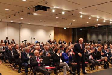 Ein großes Publikum, hauptsächlich Männer in Anzügen, bei einer Konferenz oder Veranstaltung, klatscht und applaudiert. Ein Mann im Anzug geht nach vorne.