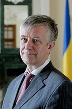 Wolodymyr Ohrysko, ehemaliger ukrainischer Außenminister, trägt Anzug und Krawatte, lächelt, mit ukrainischer Flagge im Hintergrund.