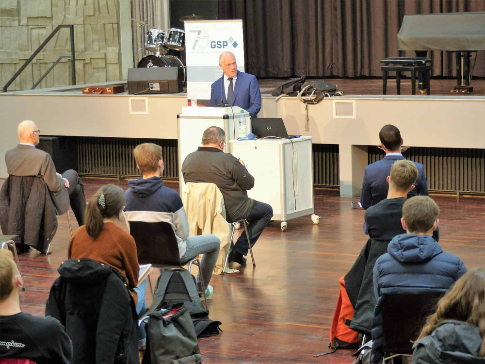 Ein Mann im blauen Anzug präsentiert von einem Pult auf einer Bühne vor einem Publikum in einem Auditorium.