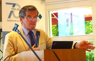 Mann mit Brille und heller Jacke spricht an einem Rednerpult, hält ein Mikrofon, mit einem „70 1952-2022“-Banner im Hintergrund.