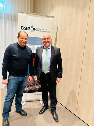 Zwei lächelnde Männer, einer in Pullover und Jeans, einer im Anzug, vor einem GSP (Gesellschaft für Sicherheitspolitik) Banner.