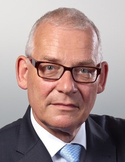 Kopfportrait eines Mannes mit grauem Haar, Brille, der einen dunklen Anzug und eine hellblaue Krawatte trägt und nach vorne blickt.