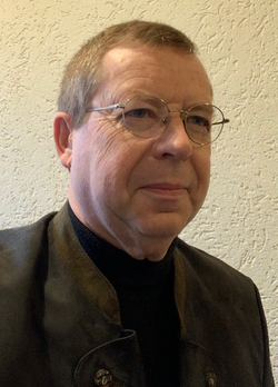 Porträt eines Mannes mit hellbraunem Haar, runder Brille und dunkler Jacke über einem schwarzen Rollkragenpullover.