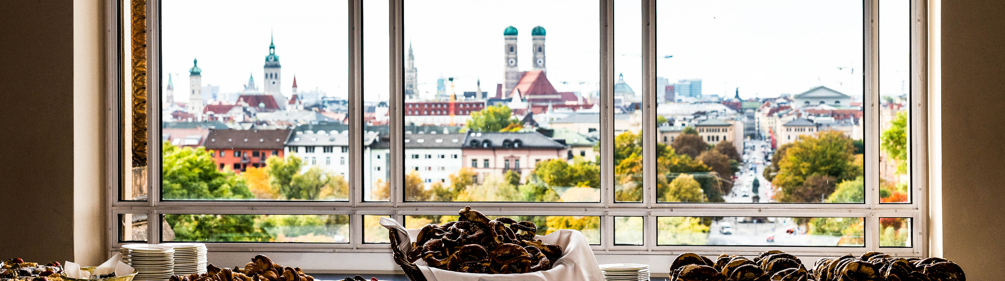 Blick aus einem Veranstaltungssaal über die Münchner Innenstadt mit den Türmen der Frauenkirche – im Vordergrund ein Buffet mit Gebäck